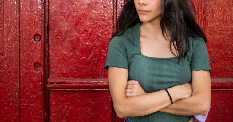 Young woman, arms crossed and looking impatient against a red door.