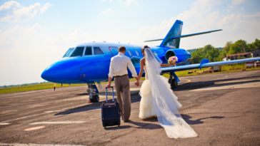A bride and a groom walking towards a plane with a suitcase.