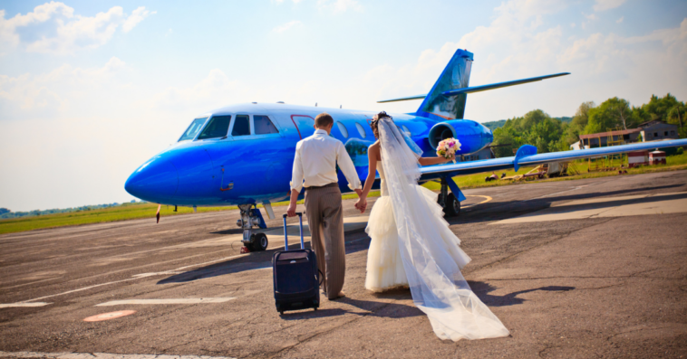 A bride and a groom walking towards a plane with a suitcase.