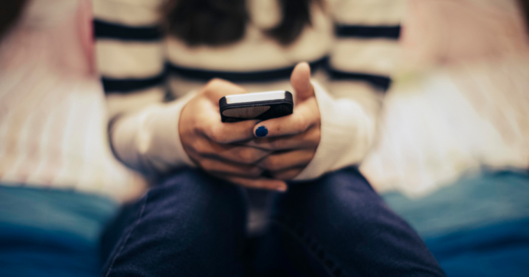 A teenage girl sitting on her bed, looking at her phone.