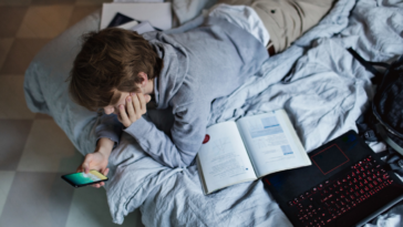 teen laying on bed with laptop and textbooks looking at phone