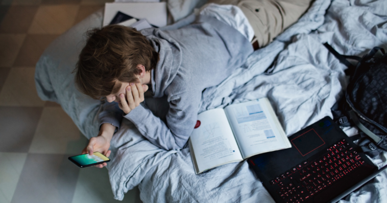 teen laying on bed with laptop and textbooks looking at phone