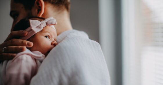 A newborn is held by her father as they stand in front of a window.