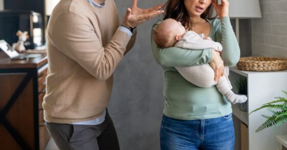 A woman rubs her temple and holds a baby as a man yells at her.