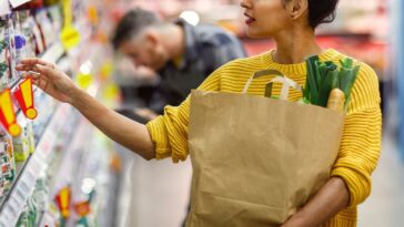 A woman, dressed in a yellow sweater, selects products in a grocery store, carrying a paper bag with fresh vegetables peeking out.