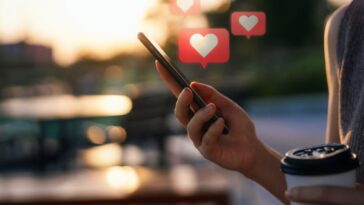 Close up of young woman checking social media network on a smartphone while holding a cup of coffee, in the city. Mobile phone with heart emoji speech bubble messages are on the screen and floating in the air.