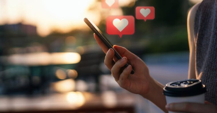 Close up of young woman checking social media network on a smartphone while holding a cup of coffee, in the city. Mobile phone with heart emoji speech bubble messages are on the screen and floating in the air.