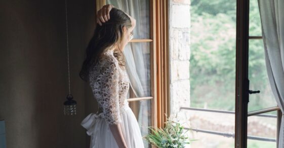 Young woman in elegant wedding dress holding bouquet looking out of the window.