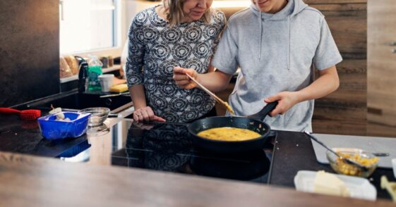 Mother and teenage son are preparing breakfast together. They are making the scrambled eggs. The boy is mixing the eggs in the frying pan.