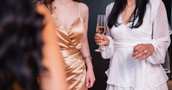 A woman in a white dress holding a glass of Champagne talking to two other women.