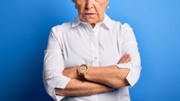 Beautiful senior age woman wearing elegant shirt standing over isolated blue background with a disapproving expression on her face and her arms are crossed.