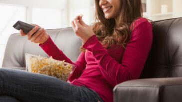 Woman watching television on sofa while eating popcorn.