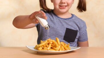 A little girl sits at a table and shakes salt on her fries.