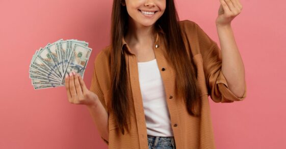 Teen Girl Holding Cash In Hands And Smiling At Camera. Standing In Front of a Pink Background.