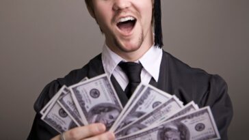 A young man, college graduate in a black cap and gown sifts through tons on money in his hands.