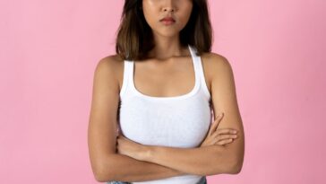 Serious young woman crossing arms and looking into camera against a plain pink background.