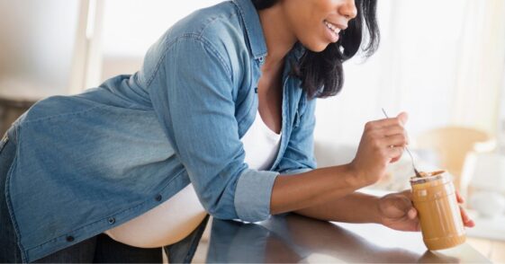 Pregnant woman leaning over a counter while eating peanut butter in the kitchen.