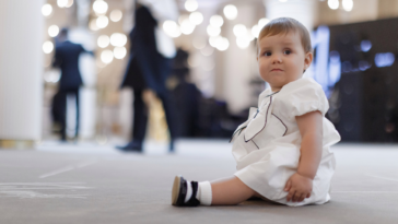 A baby in a white dress sitting on the floor at a wedding