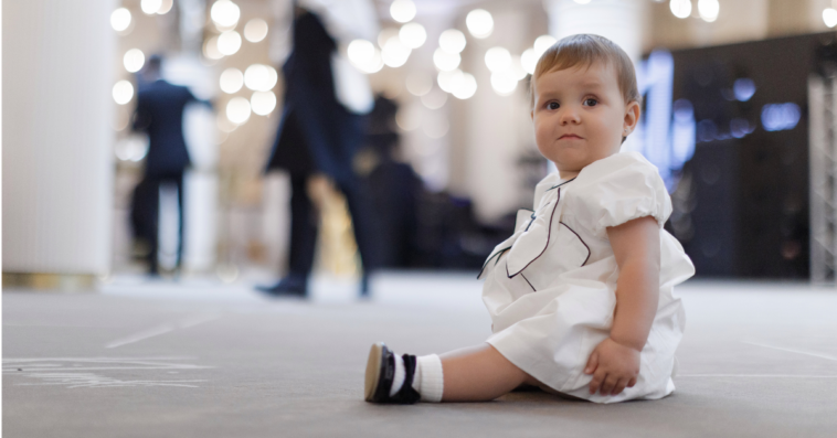 A baby in a white dress sitting on the floor at a wedding