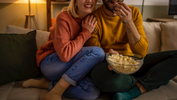 A couple on a sofa watching a movie together and eating popcorn.