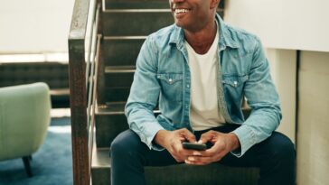 Smiling young businessman wearing glasses and reading text messages on a cellphone while sitting on stairs in an office.