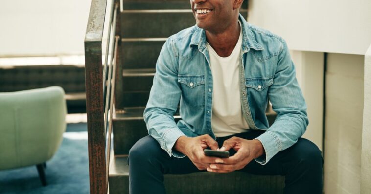 Smiling young businessman wearing glasses and reading text messages on a cellphone while sitting on stairs in an office.