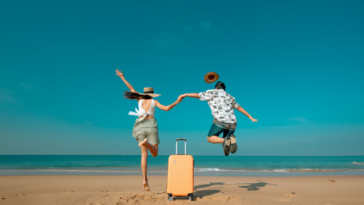 A man and a woman jumping on the beach with a suitcase behind them.