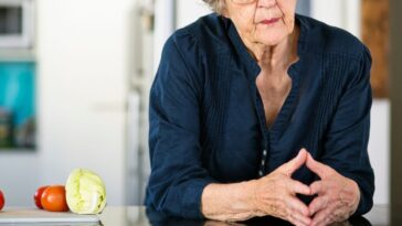 An anxious looking senior woman leans on her kitchen counter, thinking.