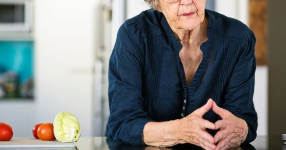 An anxious looking senior woman leans on her kitchen counter, thinking.