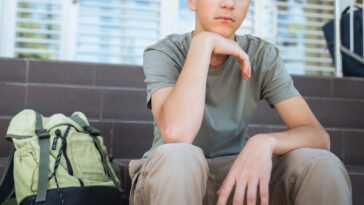 Lonely male student with a backpack on the floor sits on a staircase near school doors.