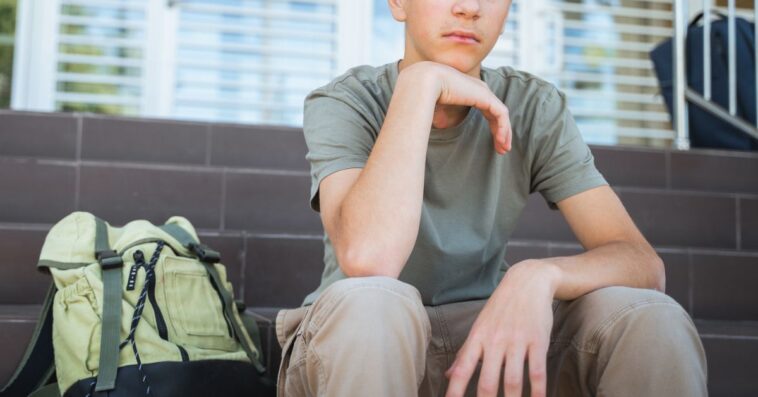 Lonely male student with a backpack on the floor sits on a staircase near school doors.