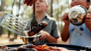 A man grilling meat on a grill with two children watching and holding plates.