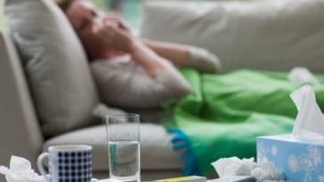 A woman lying on the sofa blowing her nose, with a tissue box, glass of water and coffee mug in front of her.