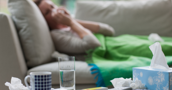 A woman lying on the sofa blowing her nose, with a tissue box, glass of water and coffee mug in front of her.