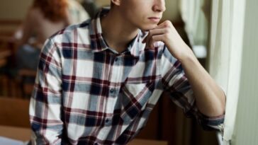 Lonely high school student, male, feeling excluded while looking through window during a break in the classroom.