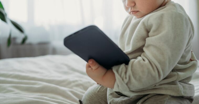 Toddler boy holding smart phone on bed at home.