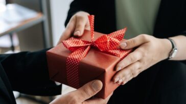 Close-up view of hands of an unrecognizable woman giving a red gift box tied with a bow. She's handing it to a man.