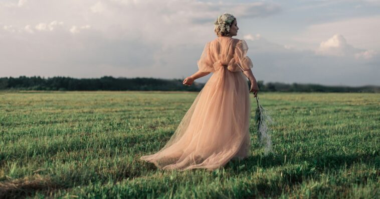 Rear view of a woman wearing vintage pink dress while walking in a field against the sky.