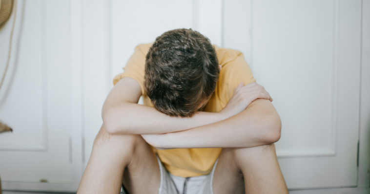 A teenage boy sitting on the floor with his head in his hands.