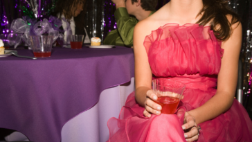 A teen girl in a pink prom dress sitting down and holding a drink.