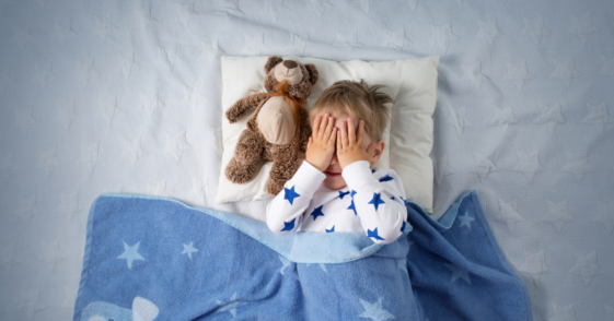 A Little boy in bed with his hands covering his eyes.