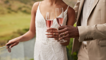 A bride and groom standing on a terrace holding glasses of chapagne.