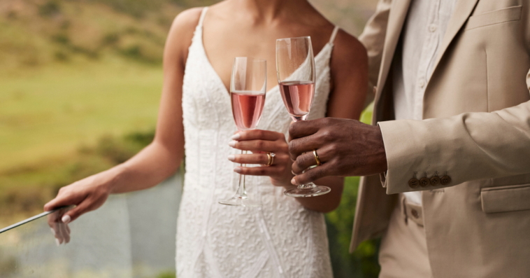 A bride and groom standing on a terrace holding glasses of chapagne.