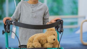 A little boy with a shaved head is smiling as he practices walking with a black walker