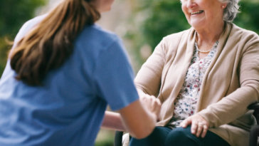 A woman in medical scrubs holding the hand and talking to an elderly woman in a wheelchair.