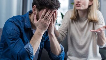 A woman shouts at her husband, in despair, crying. She requires an explanation. The man listens, covering his face with his hands. Sitting on the couch at home.