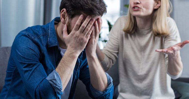 A woman shouts at her husband, in despair, crying. She requires an explanation. The man listens, covering his face with his hands. Sitting on the couch at home.