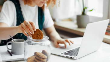 A woman at her computer dipping a cookie into a glass of milk.