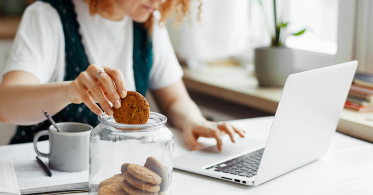 A woman at her computer dipping a cookie into a glass of milk.