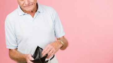 An older man standing against a pink background shows the camera an empty wallet.